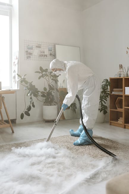 A professional cleaner wearing a white protective suit, gloves, and blue shoe covers is vacuuming a light-colored, plush carpet in a bright, minimalist living room with white walls and natural light c